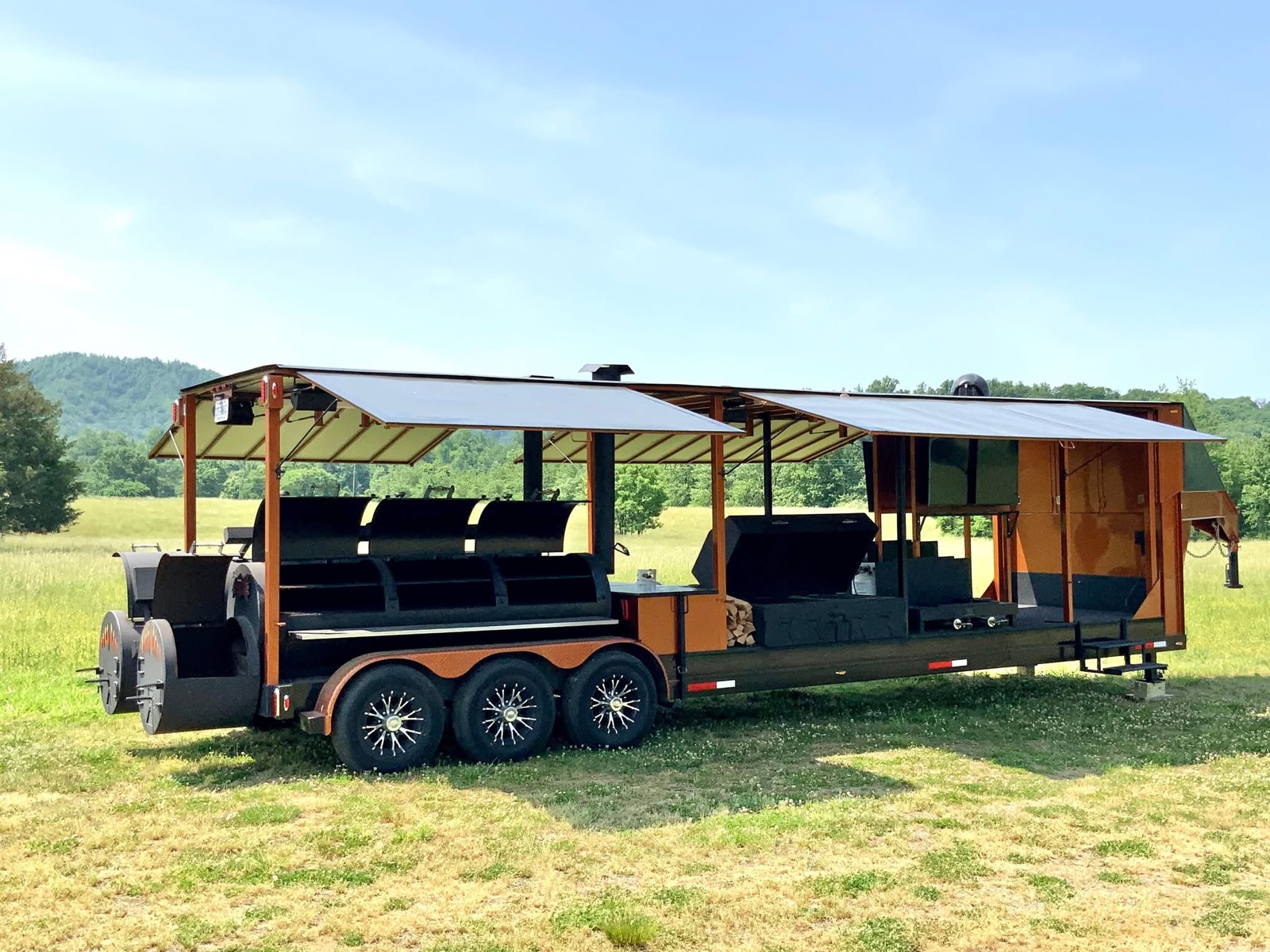 The BBQ Beast rig — an orange gooseneck trailer with a triple-barrel smoker under its own canopy, parked in a Blue Ridge meadow