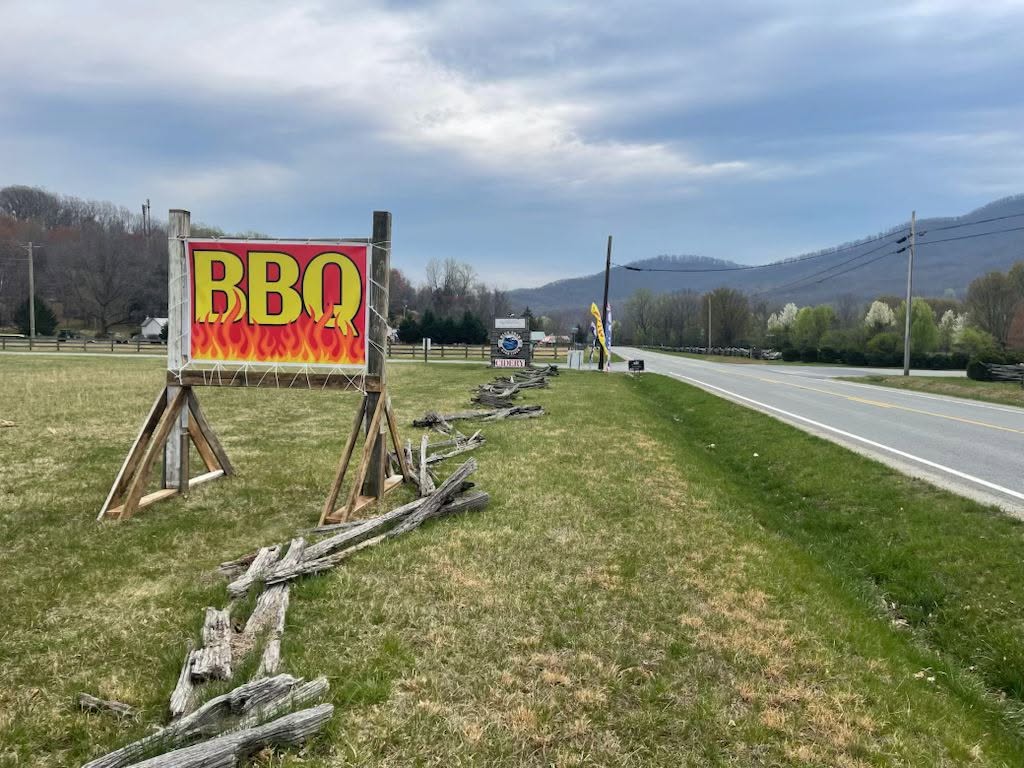 A hand-built roadside sign reading BBQ in flames, mountains down the road behind it